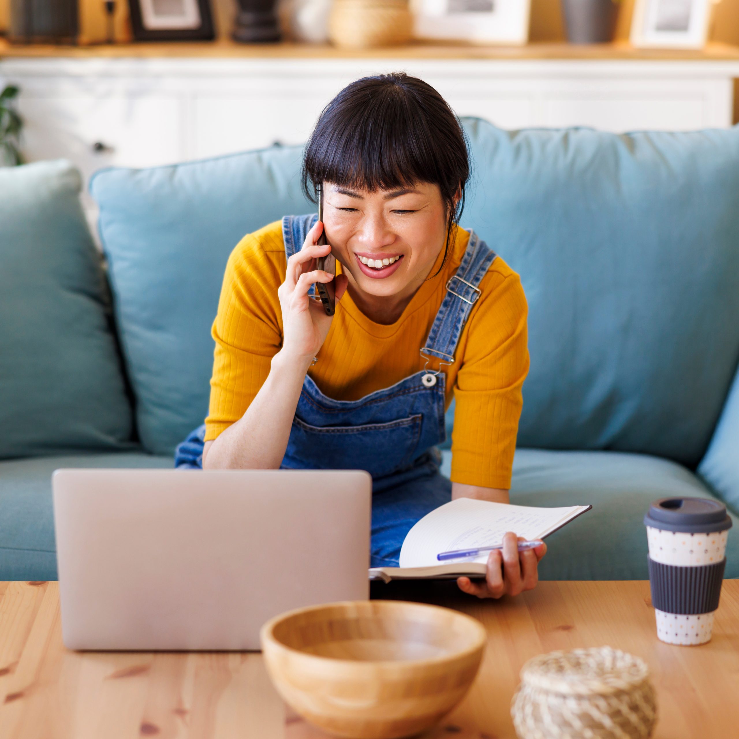 A woman work while seated on the couch, talking on the phone,  smiling at her laptop, and holding a notebook